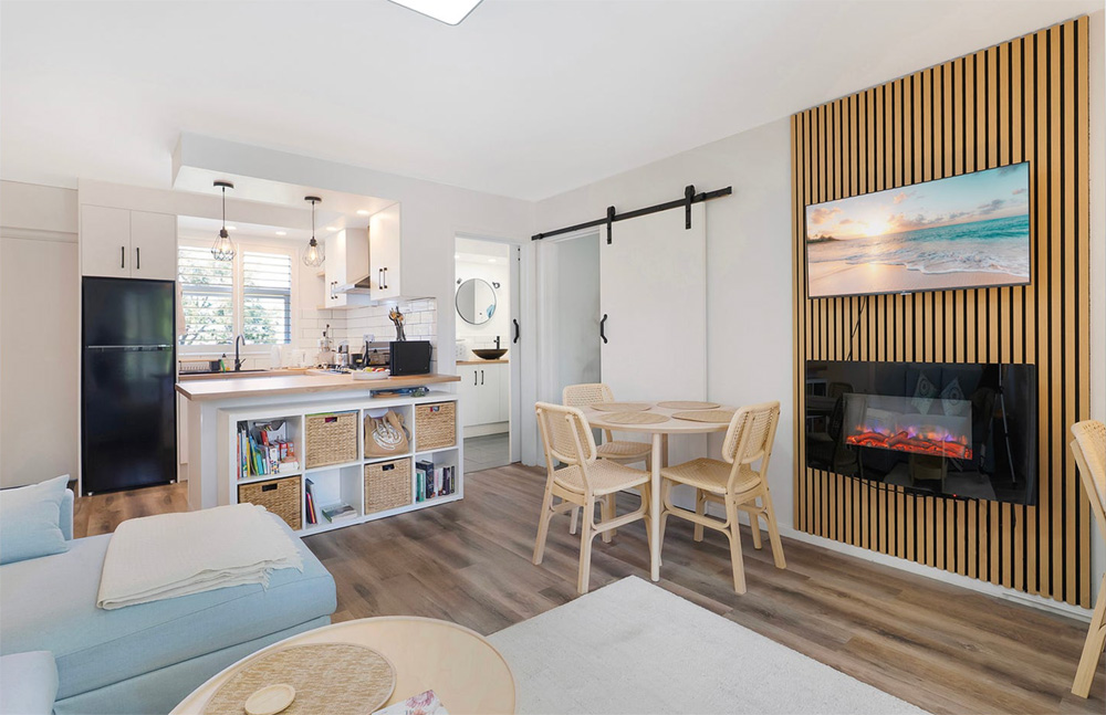 Wide angle photo of a clean and bright apartment living room with natural timber bench tops and white cupboards.
